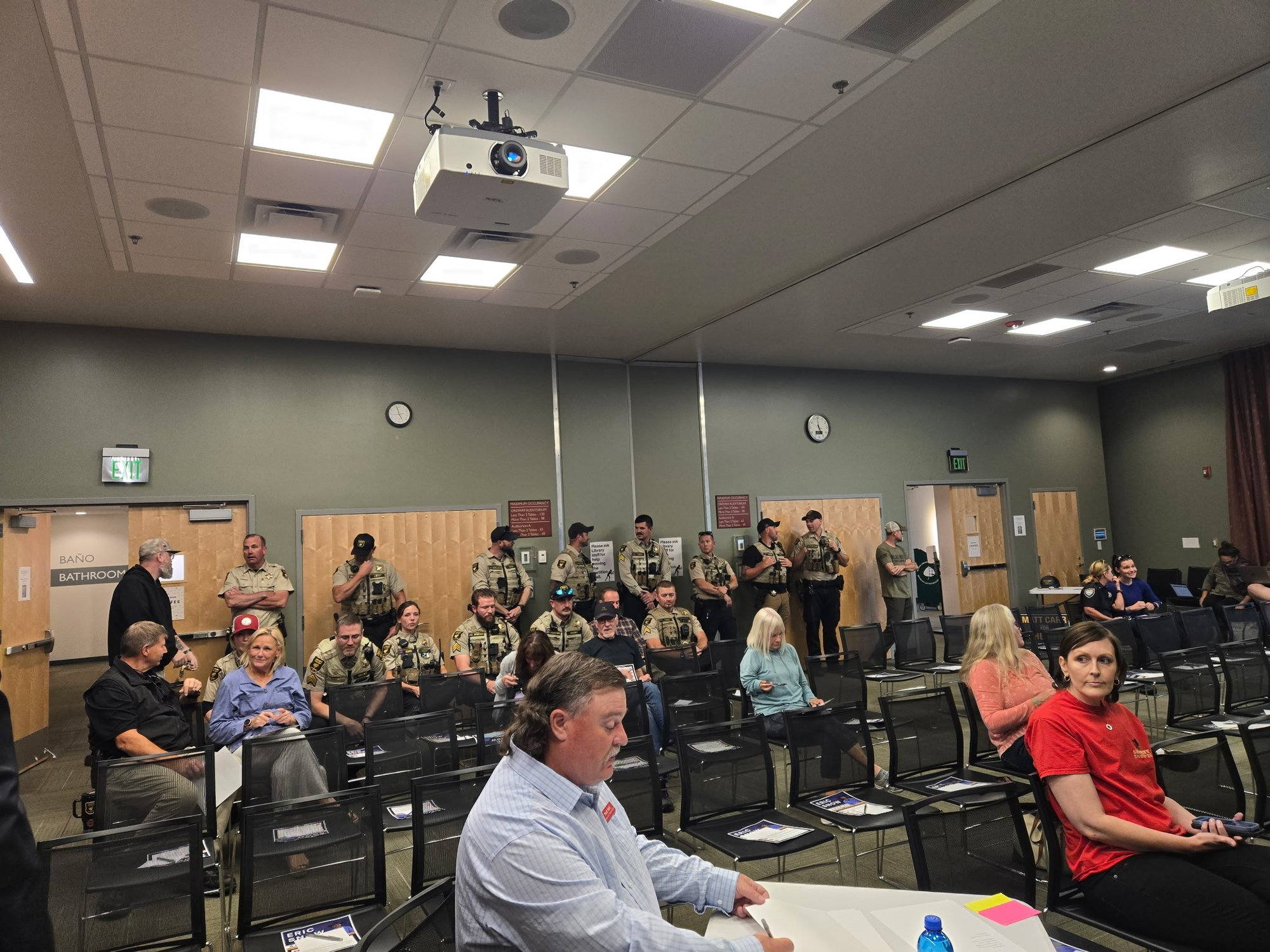 The back of the library meeting room at the October 5 forum: about ten deputies in tan duty uniform with vests, radios, and sidearms standing along the wall, and several more seated, among civilian attendees holding Eric Snow flyers.