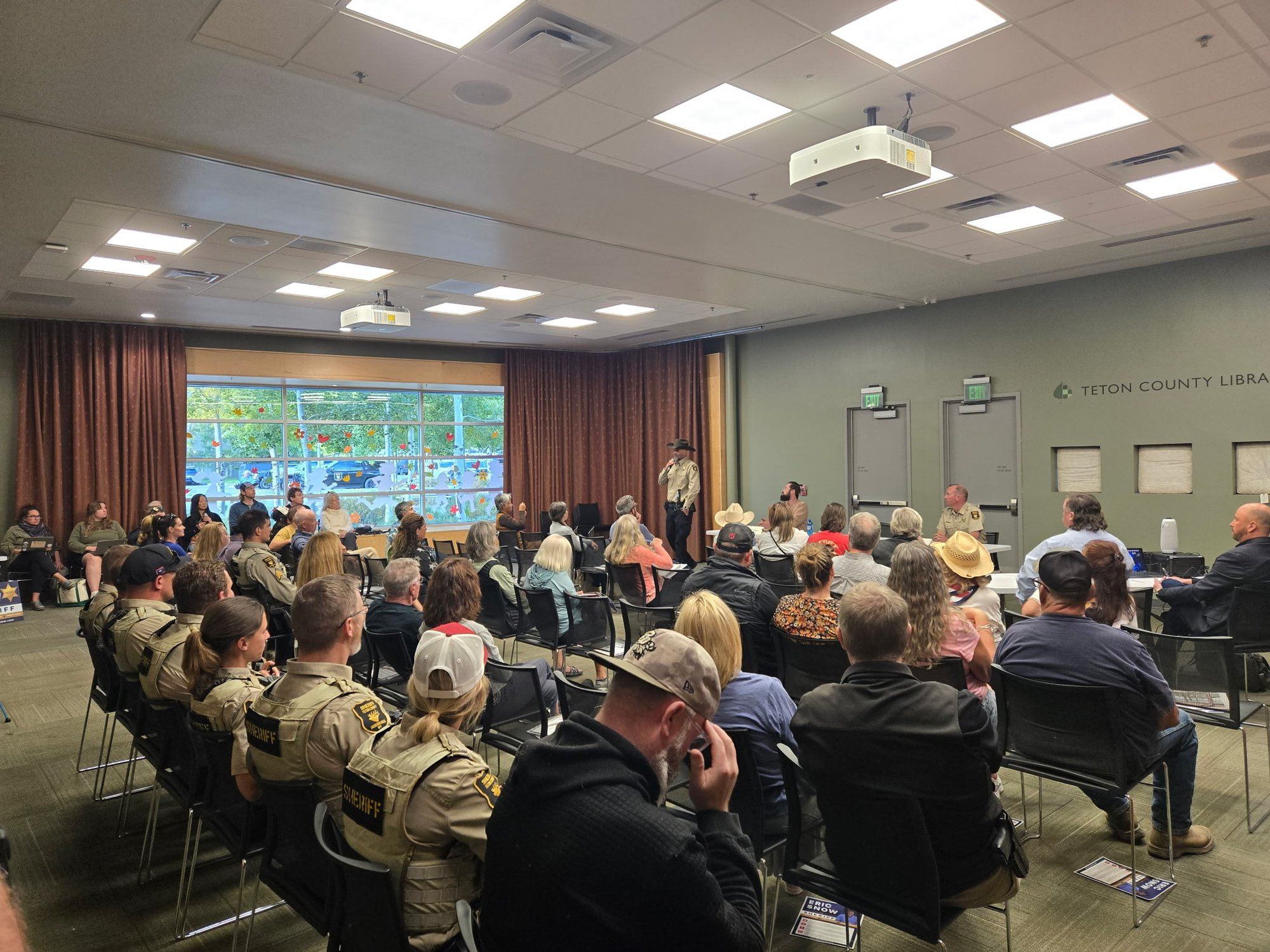 Deputy Eric Snow in uniform and cowboy hat speaking into a microphone at the front of the room while four deputies in SHERIFF vests sit together in the left rows; Sheriff Carr in uniform seated at the end of the front table.