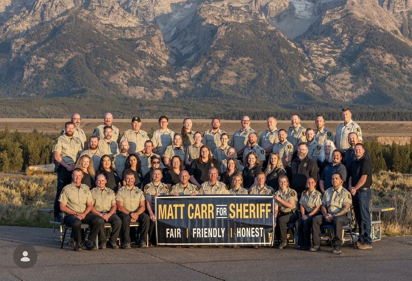 Roughly 45 Teton County Sheriff's Office personnel in County uniform, in rows in front of the Tetons, holding a Matt Carr for Sheriff banner.