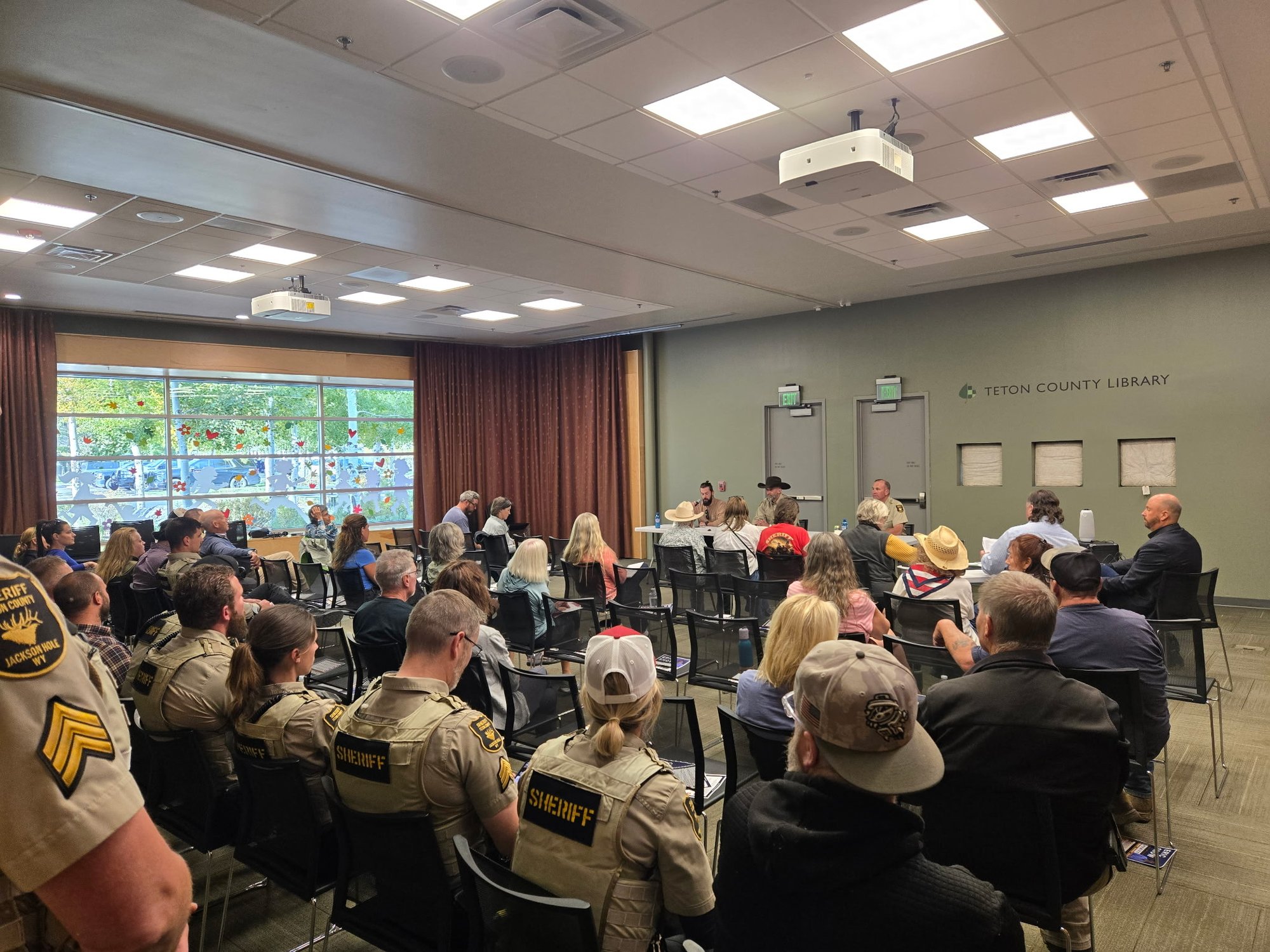 A sergeant's sleeve with Teton County Sheriff patch and chevrons in the foreground; seated deputies in SHERIFF vests; the moderator and the candidates, Carr and Snow in uniform, at the front table.
